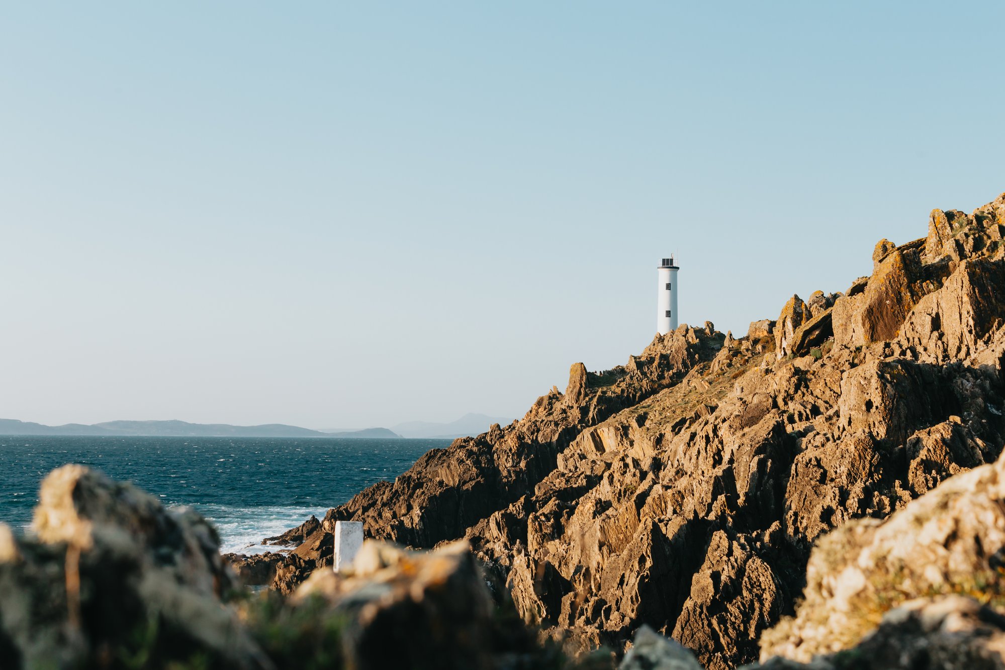tall lighthouse on sharp rocky shoreline against blue water 高高的灯塔，在陡峭的岩石海岸线上，靠蓝色的海水。