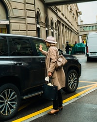 man in brown coat and black hat standing beside black suv during daytime 白天，身穿棕色外套，戴着黑色帽子的男子站在黑色SUV旁边。