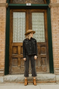 woman in black leather jacket and brown hat standing in front of brown wooden door 身穿黑色皮夹克和棕色帽子的女子站在棕色木门前