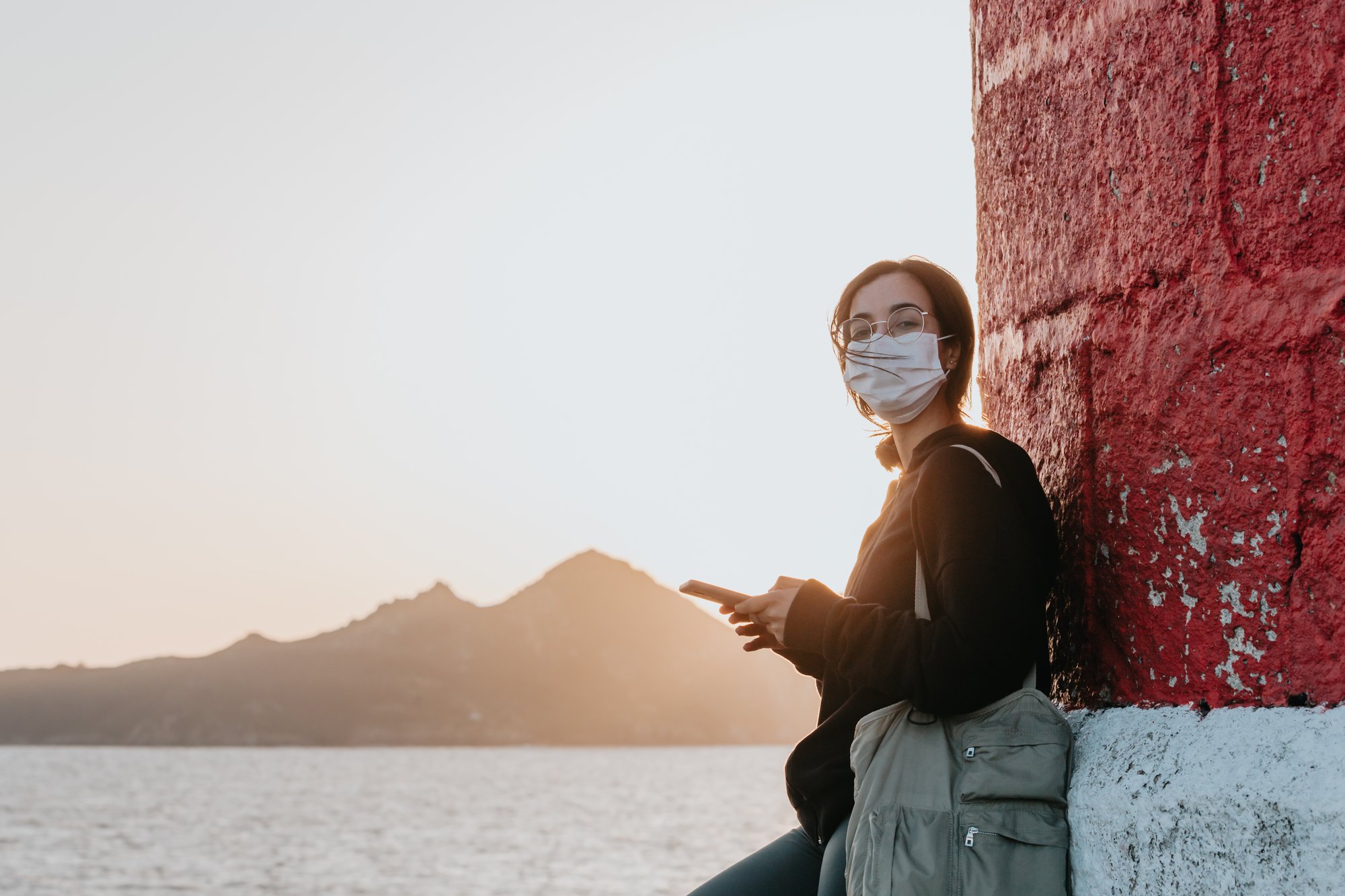 woman in facemask leans against a wall by water 戴面罩的女人靠在水边的墙上