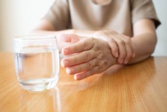 老年妇女的手颤抖着症状伸出来在木桌上取一杯水。握手的原因包括帕金森病、中风或脑损伤。精神健康神经紊乱
