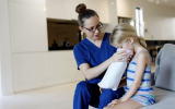 A healthcare provider holds a disposable vomit bag steadily for the young blonde girl beside her and helps her carefully.