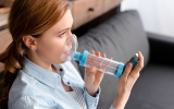A young woman sits comfortably on a sofa at home, holding an inhaler spacer with a mask to her face to inhale respiratory medication calmly.